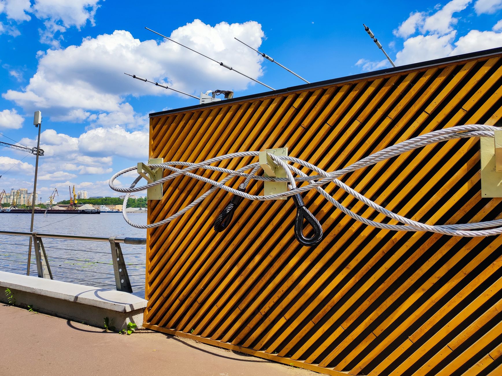Diagonal striped wall with attached ropes by riverside, blue sky with clouds.